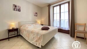 Un dormitorio con cama, mesitas de noche, una silla y un ventanal con cortinas.