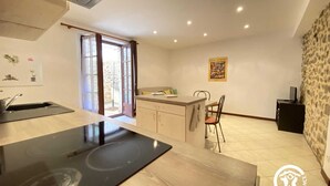 Una cocina con estufa, lavamanos y zona de comedor. Hay una ventana con cortinas, una pared de piedra y una mesa pequeña con sillas.