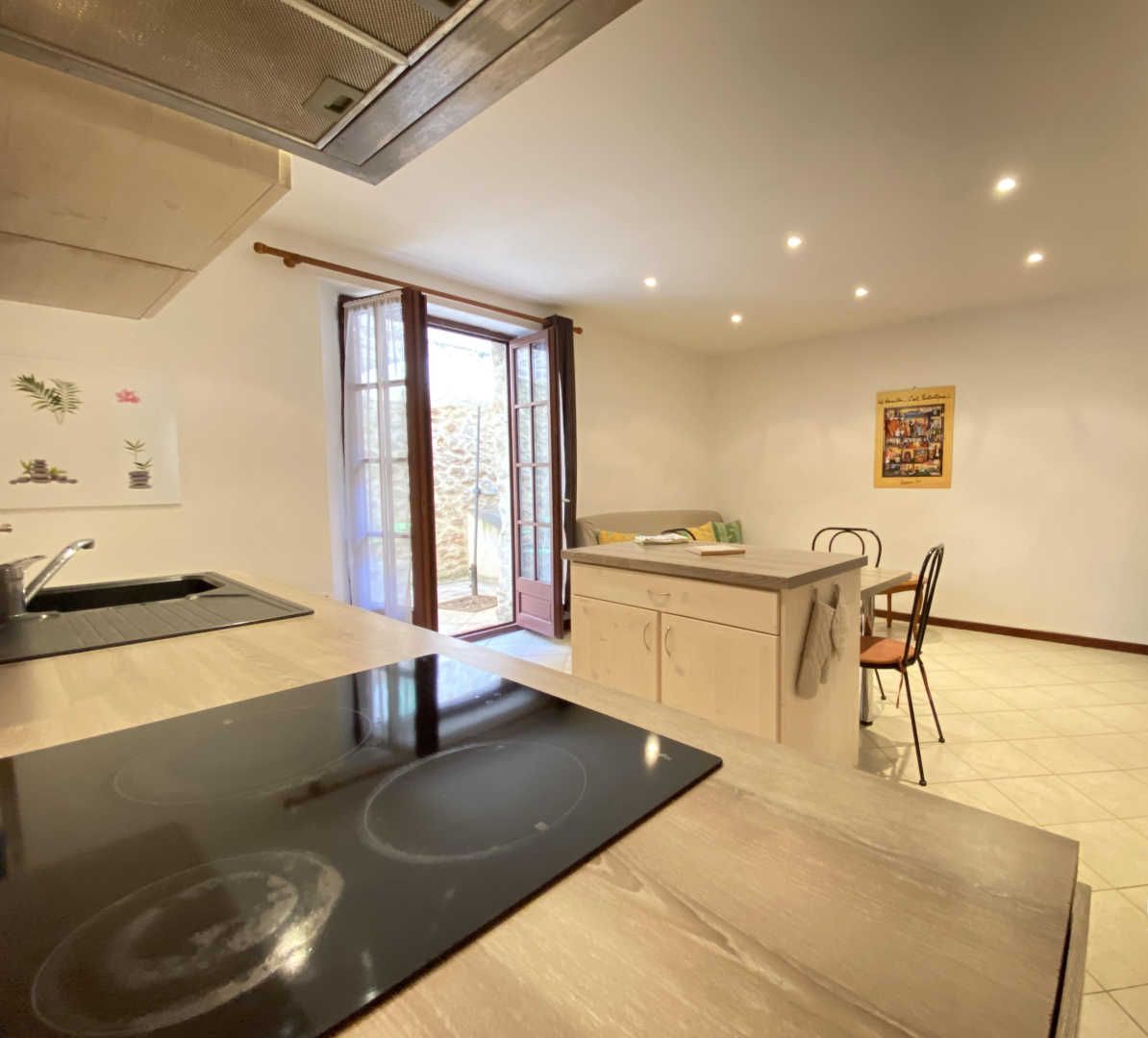 A kitchen with a stove, sink, and dining area. There is a window with curtains, a stone wall, and a small table with chairs.