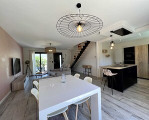 Una cocina moderna con una isla central, un comedor con mesa blanca y una escalera que lleva a un nivel superior.