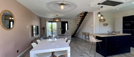 A modern kitchen with a central island, a dining area with a white table, and a staircase leading to an upper level.