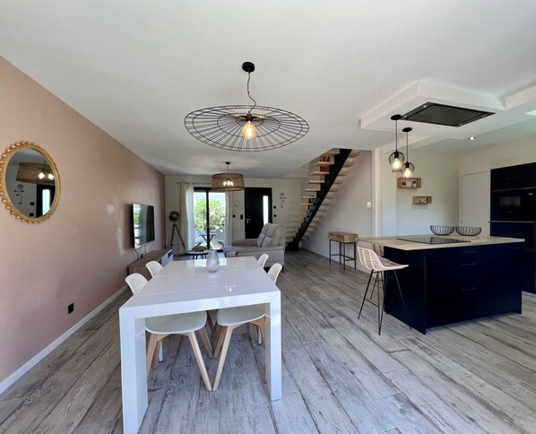Una cocina moderna con una isla central, un comedor con mesa blanca y una escalera que sube a un nivel superior.