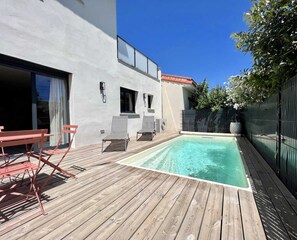Una terraza de madera con piscina, mobiliario exterior y un edificio blanco con ventanas y una puerta.
