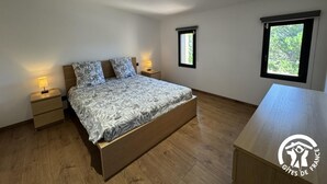 Une chambre à coucher avec un lit en bois, deux tables de chevet avec des lampes, une petite fenêtre et une plus grande fenêtre donnant sur un paysage verdoyant.