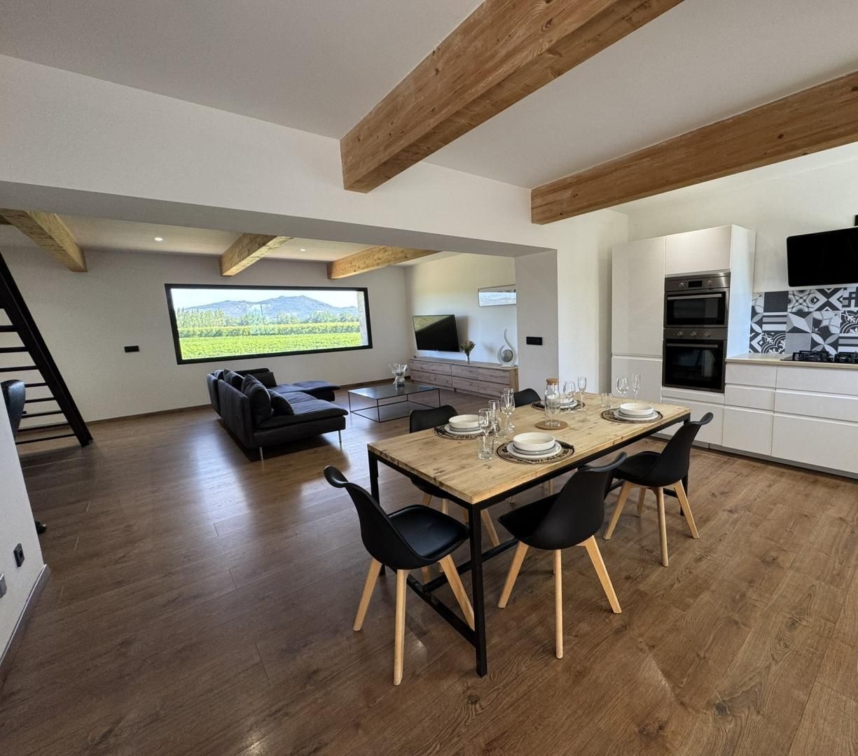 Een moderne keuken met witte kasten, een houten eettafel en zwarte stoelen. Er is een televisie aan de muur bevestigd en een raam met uitzicht op een groen landschap.