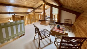 A cozy attic room with wooden beams, a balcony with a railing, and a seating area with chairs and a table.