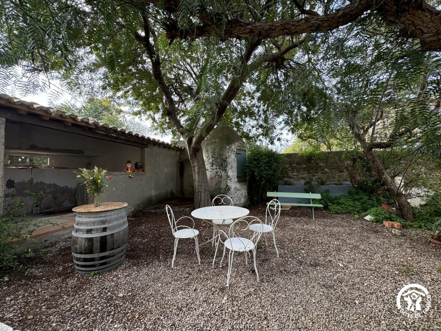 Een terras met een ronde tafel en vier stoelen, omringd door grind en een grote boom.