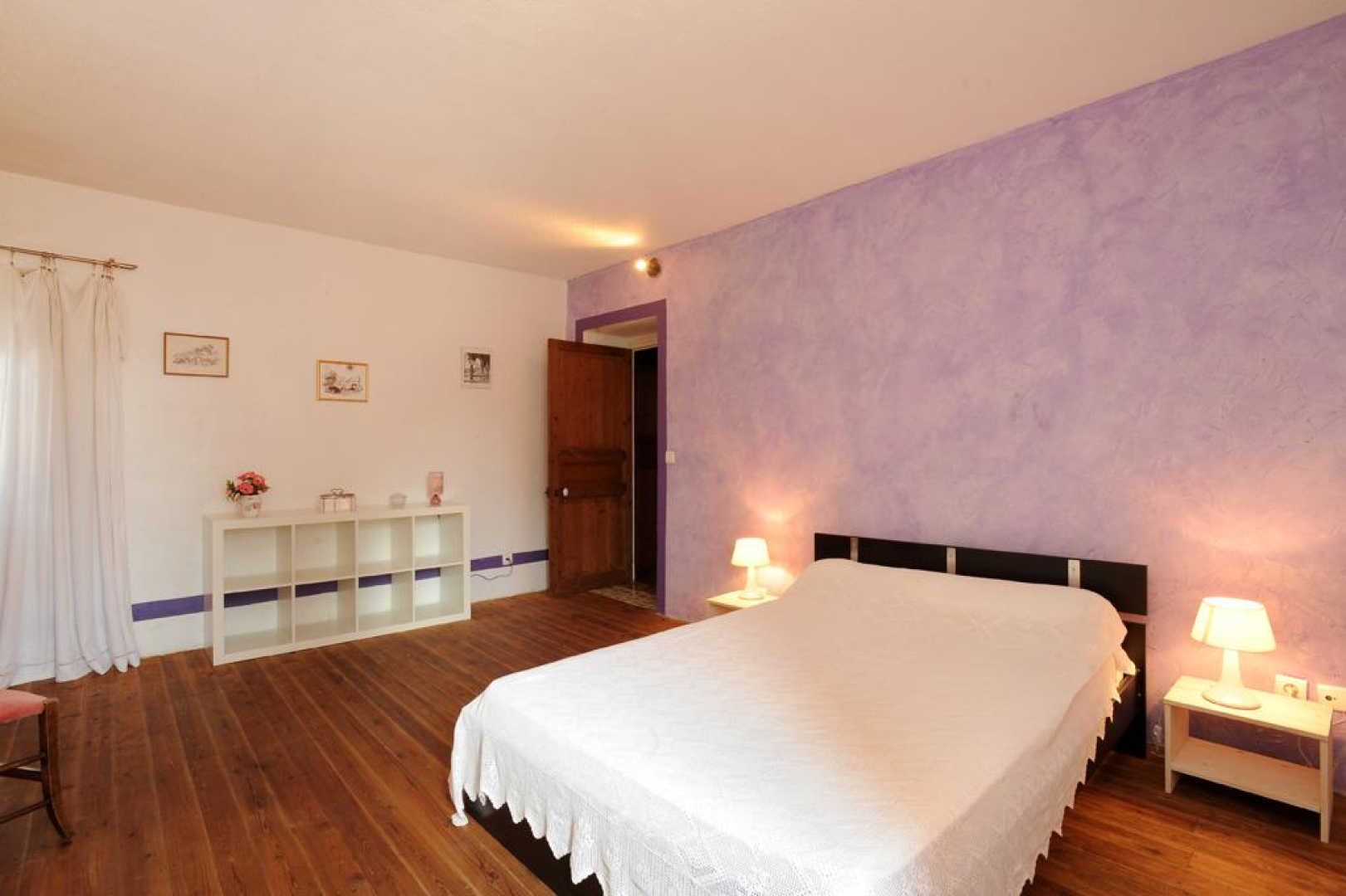 A bedroom with a large bed, a nightstand with a lamp, a wooden dresser, and a door leading to another room.