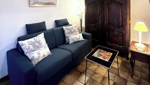 A living room with a dark blue sofa, two patterned cushions, a small table with a magazine, a wooden cabinet, and a lamp on a side table. - Le Plô du Moulin - VILLENEUVE MINERVOIS (VILLENEUVE MINERVOIS)