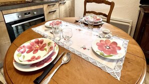A dining area with a round wooden table set for two, featuring floral tablecloths and plates. A window with curtains is visible, and a kitchen area with wooden cabinets and a stove is in the background. - Le Plô du Moulin - VILLENEUVE MINERVOIS (VILLENEUVE MINERVOIS)