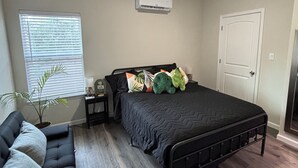 Un dormitorio con cama, ventilador de techo, aire acondicionado, ventana con persianas, una planta en maceta y un sofá de color oscuro.