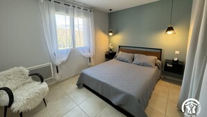 Ein Schlafzimmer mit Bett, Stuhl, Nachttisch, Lampe und einem Fenster mit Vorhängen.