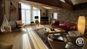 Un salon chaleureux avec un mur en pierre, des poutres apparentes, un canapé, une télévision et un fauteuil suspendu.