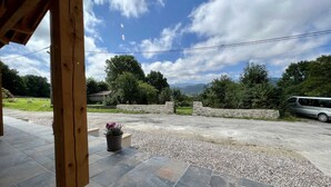 A covered outdoor area with a stone pathway leading to a vehicle, surrounded by greenery and a mountainous backdrop.
