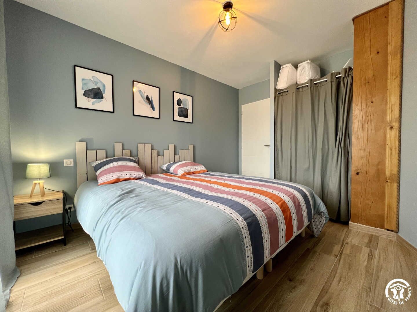 Een slaapkamer met een bed, nachtkastje, lamp en houten hoofdeinde. Er hangen drie ingelijste foto's aan de muur.