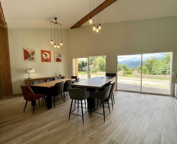 A spacious dining area with a wooden table and chairs, a ceiling light fixture, and a view of a garden through large sliding doors. - Gîte du Tatou - ROQUEFEUIL (ROQUEFEUIL)