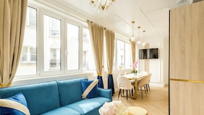 Een moderne woonkamer met een blauwe bank, een eethoek met een tafel en stoelen, en een groot raam met gordijnen.