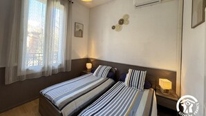 A bedroom with two beds, a window with curtains, a wall-mounted air conditioner, and a small table with a lamp.