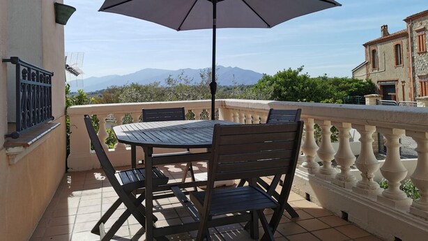 A balcony with a table and chairs, a large umbrella, and a view of mountains.