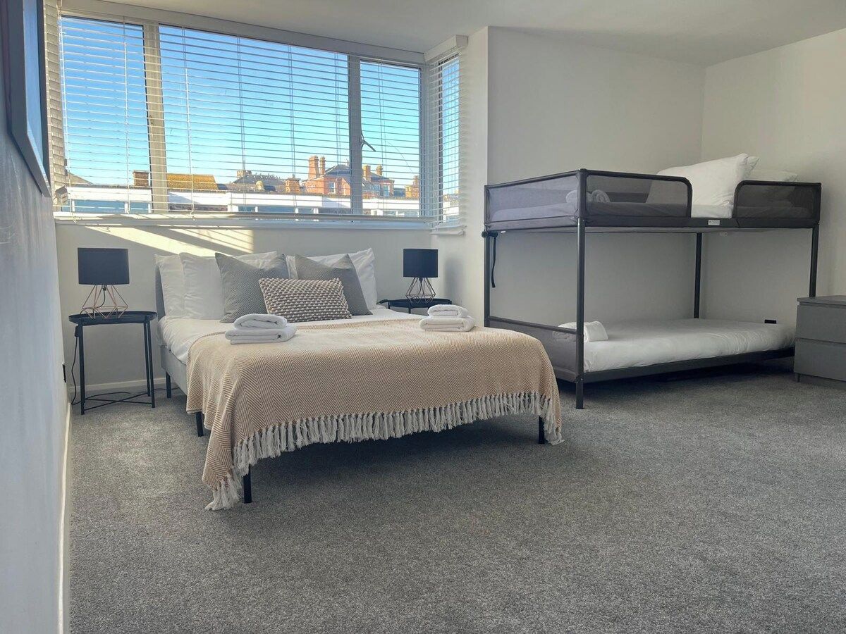 A bedroom with a bunk bed, a single bed, a lamp, and a view of the city through large windows.