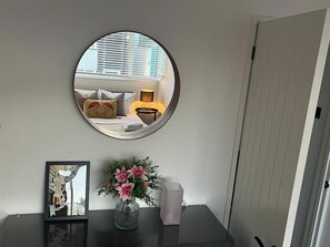 Un espejo redondo en la pared, un cuadro enmarcado, un florero con flores y una mesita con una lámpara.