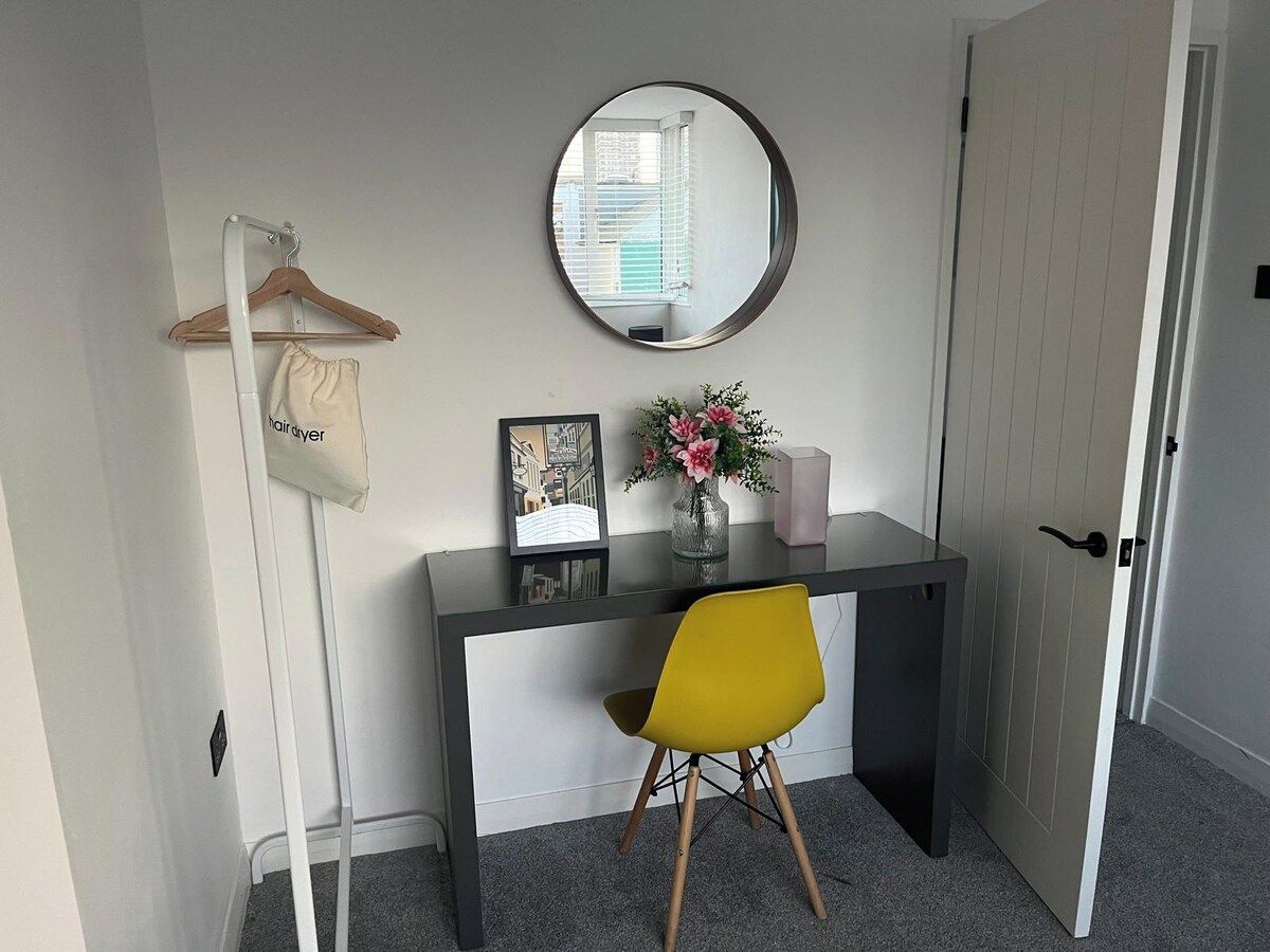 A room with a desk, a chair, a mirror, a hanging coat, and a door.