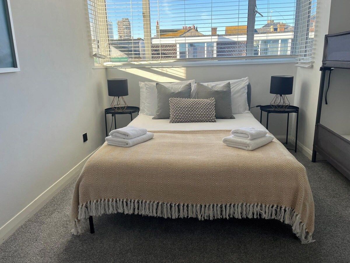 A neatly made bed with a beige and brown patterned bedspread, two pillows, and two folded towels on either side. There are two bedside tables with lamps, and the room has a large window with blinds.