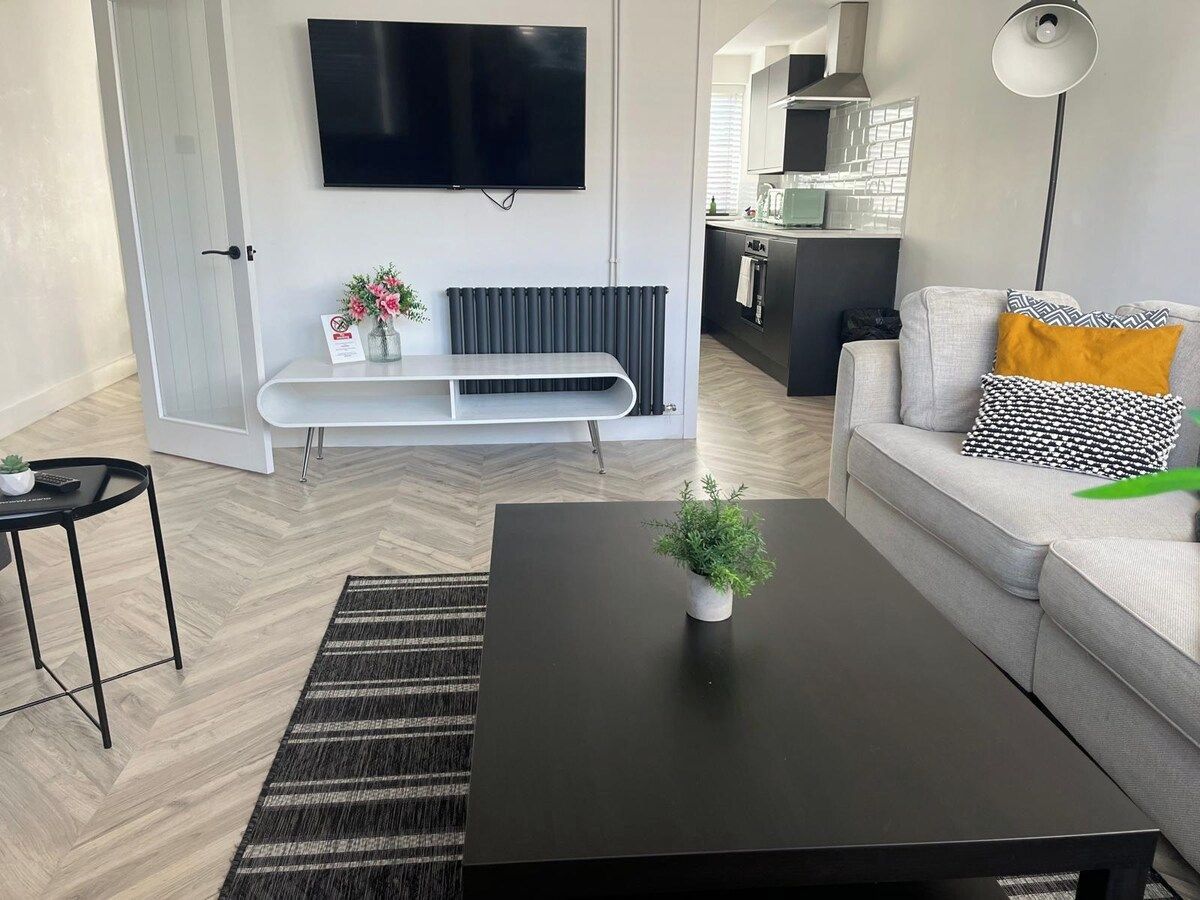 A modern living room with a black coffee table, a grey sofa with patterned cushions, a flat-screen TV mounted on the wall, and a small plant on the table.