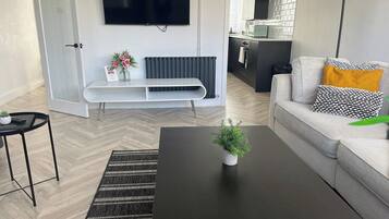 A modern living room with a black coffee table, a grey sofa with patterned cushions, a flat-screen TV mounted on the wall, and a small plant on the table.