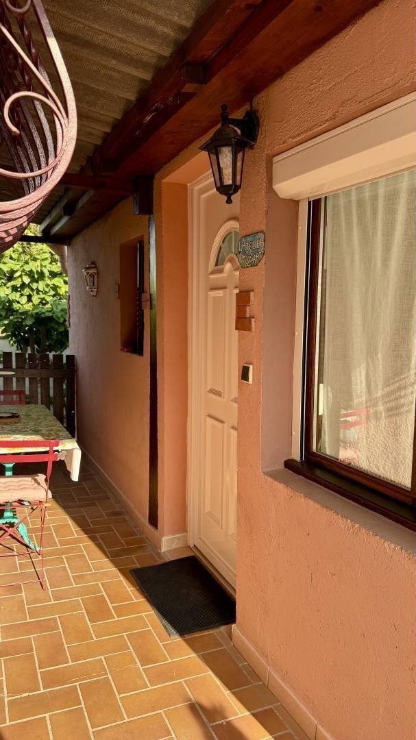 Un espace extérieur abrité, avec une table et des chaises en bois, une porte avec une enseigne et une fenêtre avec un rideau.