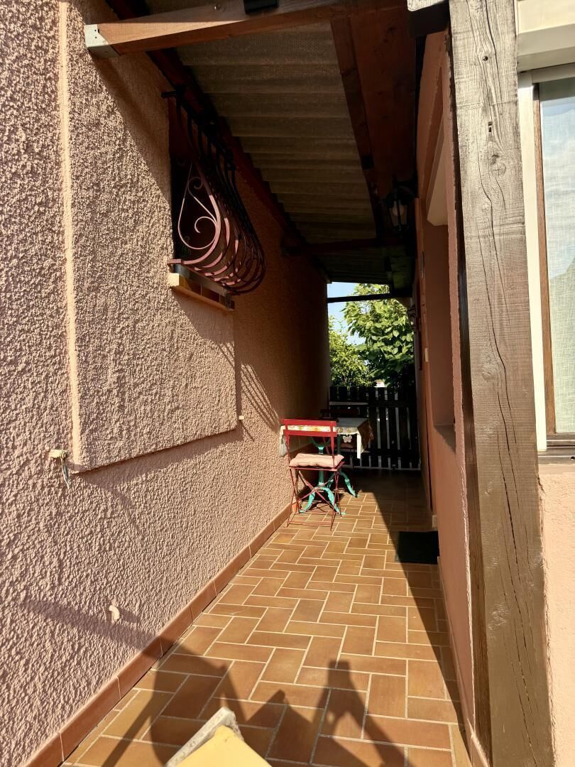 A narrow outdoor corridor with a tiled floor, a red table with chairs, and a black fence in the background.
