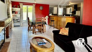 A living room with a black sofa, wooden coffee table, and a dining area with a wooden table and chairs. A kitchen with white cabinets and a wooden bar counter is visible in the background. - Le Ligno - QUILLAN - Gîte (QUILLAN)