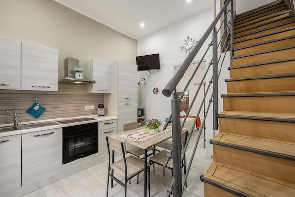 A modern kitchen with a dining area, wooden staircase, and a flat-screen TV mounted on the wall.