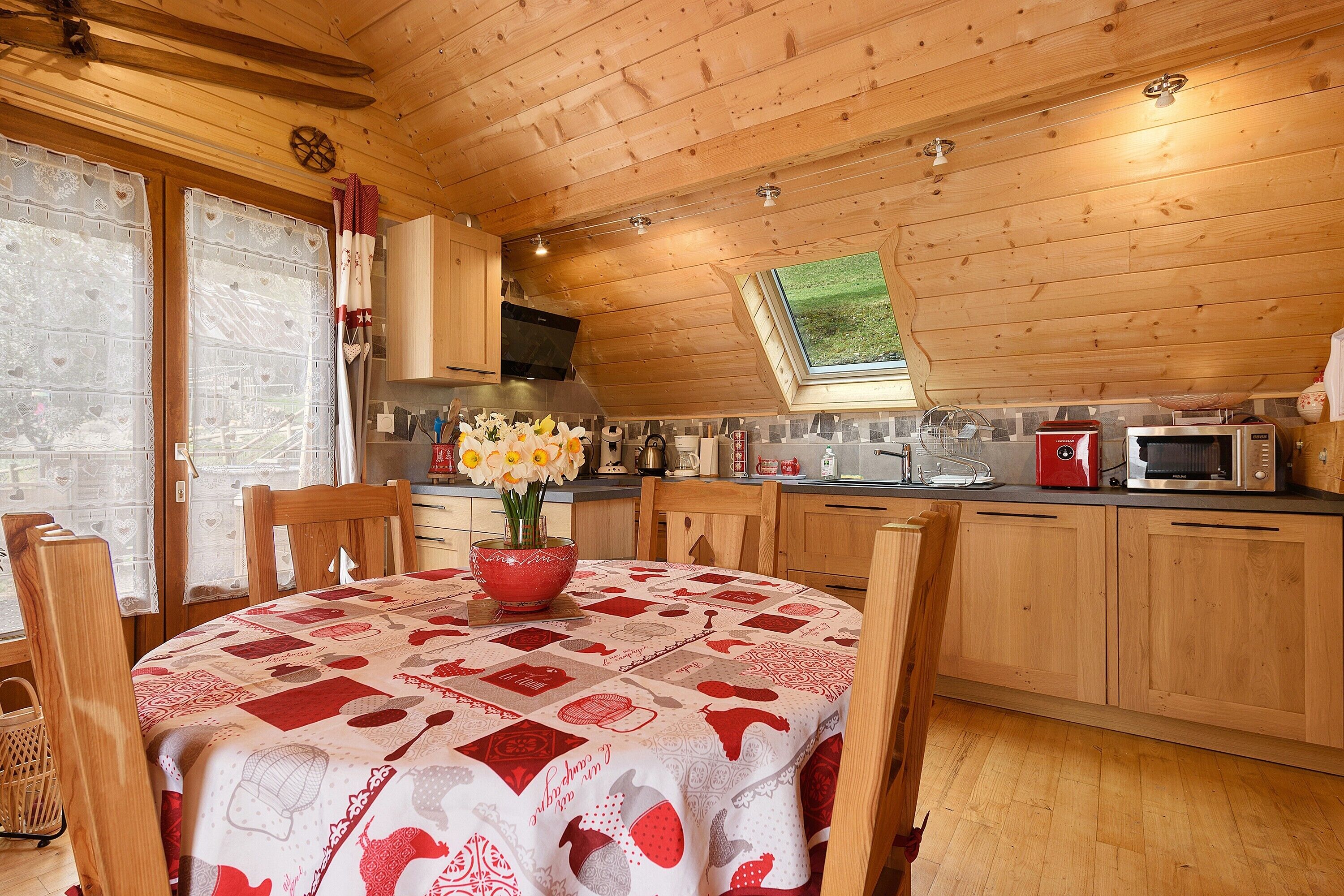 Une cuisine chaleureuse avec des meubles en bois, une table à manger recouverte d’une nappe rouge et blanche, et une fenêtre donnant sur un paysage verdoyant.
