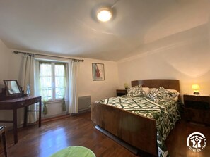 Een slaapkamer met een houten bed, een bureau en een stoel. Er is een raam met gordijnen, een schilderij aan de muur en een lamp op het nachtkastje.