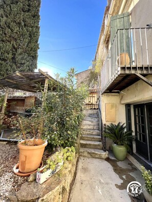 Ein schmaler Innenhof mit einer Steintreppe, die zu einem Balkon führt, umgeben von Pflanzkübeln und einem großen Baum.