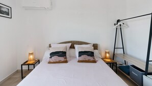 Een net opgemaakt bed met twee kussens, een bedlampje, een klein tafeltje, een kledingrek en een wandairconditioner.