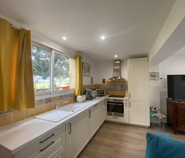 A modern kitchen with white cabinets, a microwave, an oven, and a sink. There is a window with yellow curtains, a television, and a dining area with a table and chairs.