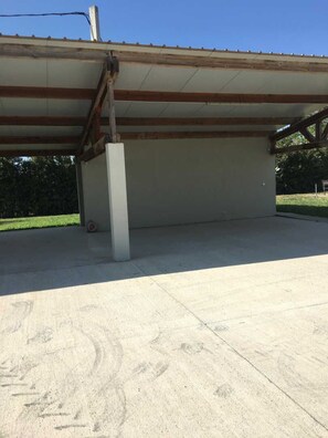 A covered outdoor area with a concrete floor, a metal roof, and a wooden beam structure. - Illibéris 2 gardens (ELNE)