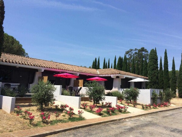 Un edificio con tejado de tejas, mobiliario exterior con sombrillas rojas y un jardín ajardinado con flores rosadas.