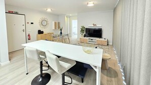 Un salon moderne avec une table à manger blanche, deux chaises, un téléviseur et une petite plante.