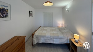 A bedroom with a bed, a wooden dresser, a nightstand with a lamp, a ceiling light, a door, and a framed picture on the wall. - Terrace - MONTALBA-LE-CHATEAU (MONTALBA-LE-CHATEAU)