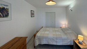 A bedroom with a bed, a wooden dresser, a nightstand with a lamp, a ceiling light, a door, and a framed picture on the wall.