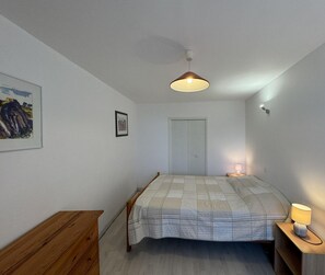 Une chambre à coucher avec un lit, une commode en bois, une table de chevet avec une lampe, un luminaire au plafond, une porte et un tableau encadré au mur.