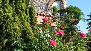 Un bâtiment en pierre, doté d’un toit en tuiles et de fenêtres en arc de cercle, entouré d’une végétation luxuriante et de rosiers roses en pleine floraison.