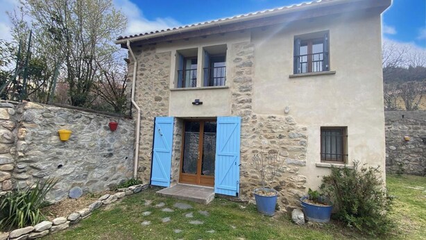 Ein zweistöckiges Steinhaus mit einer Steinmauer, einer Holztür mit blauen Fensterläden und einem kleinen Vorgarten mit Blumentöpfen.