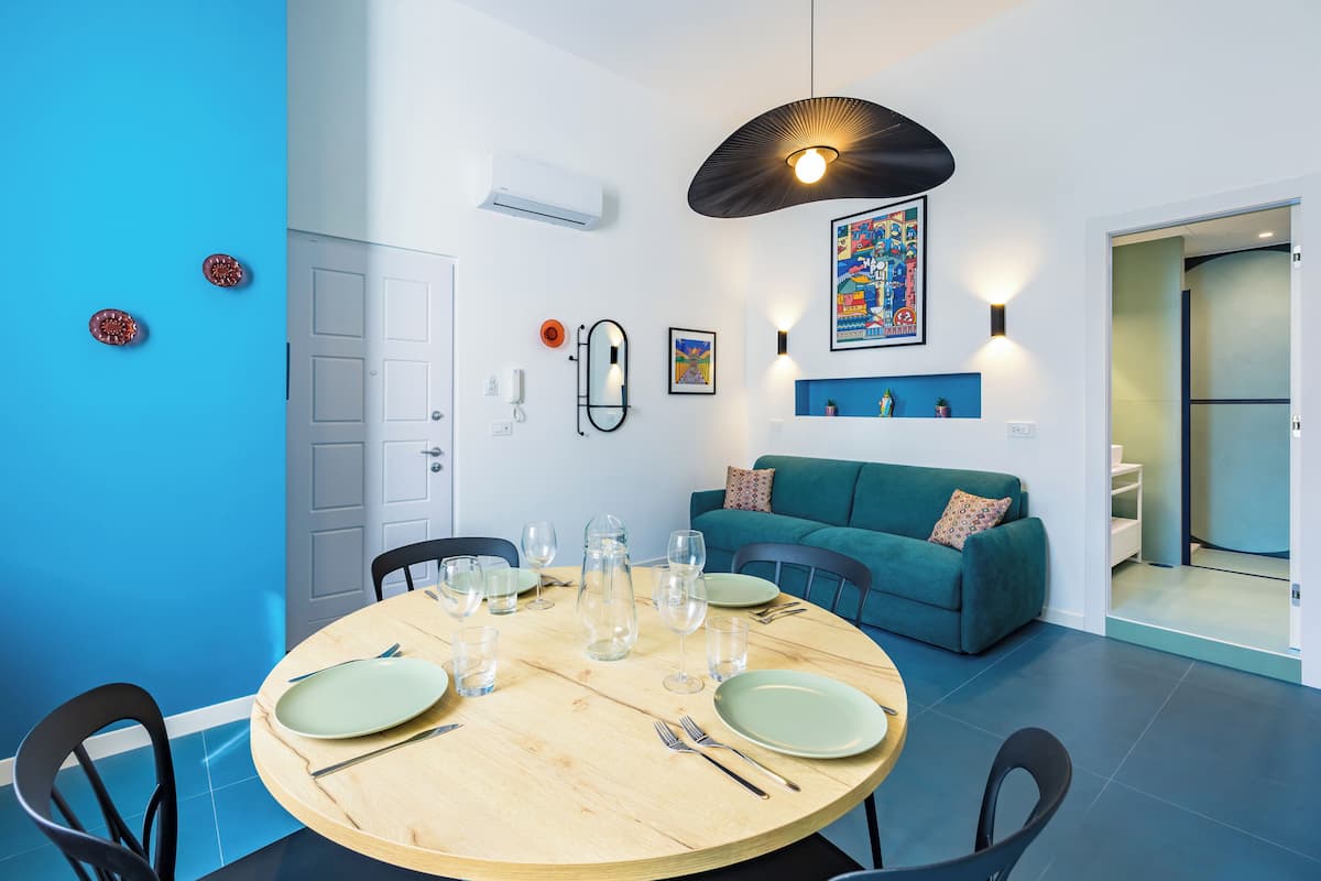 A dining area with a round wooden table set for four, surrounded by black chairs. A teal sofa is against the wall, and a colorful abstract painting is above it.