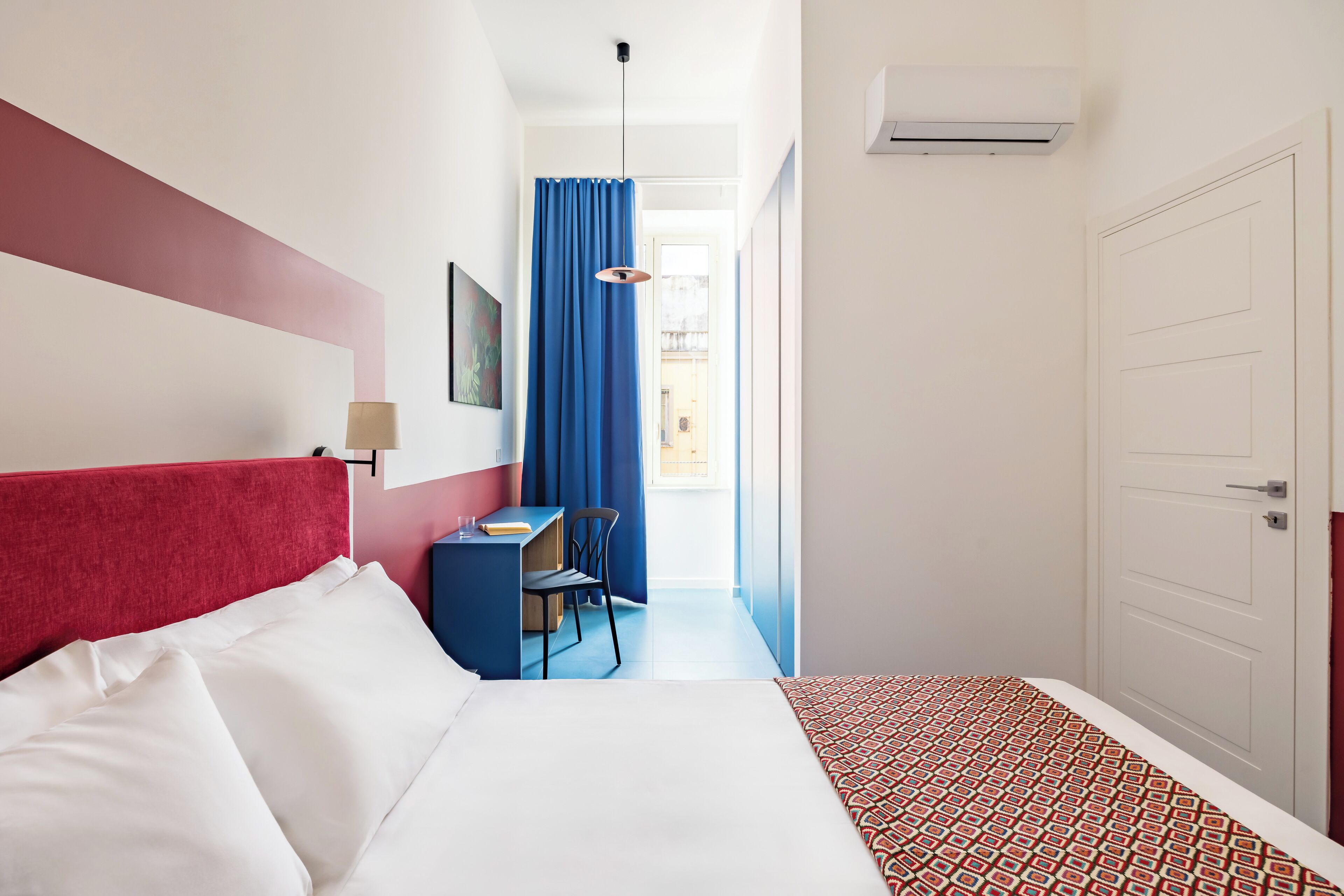 A hotel room with a bed, a desk, a chair, a blue curtain, and a white door.