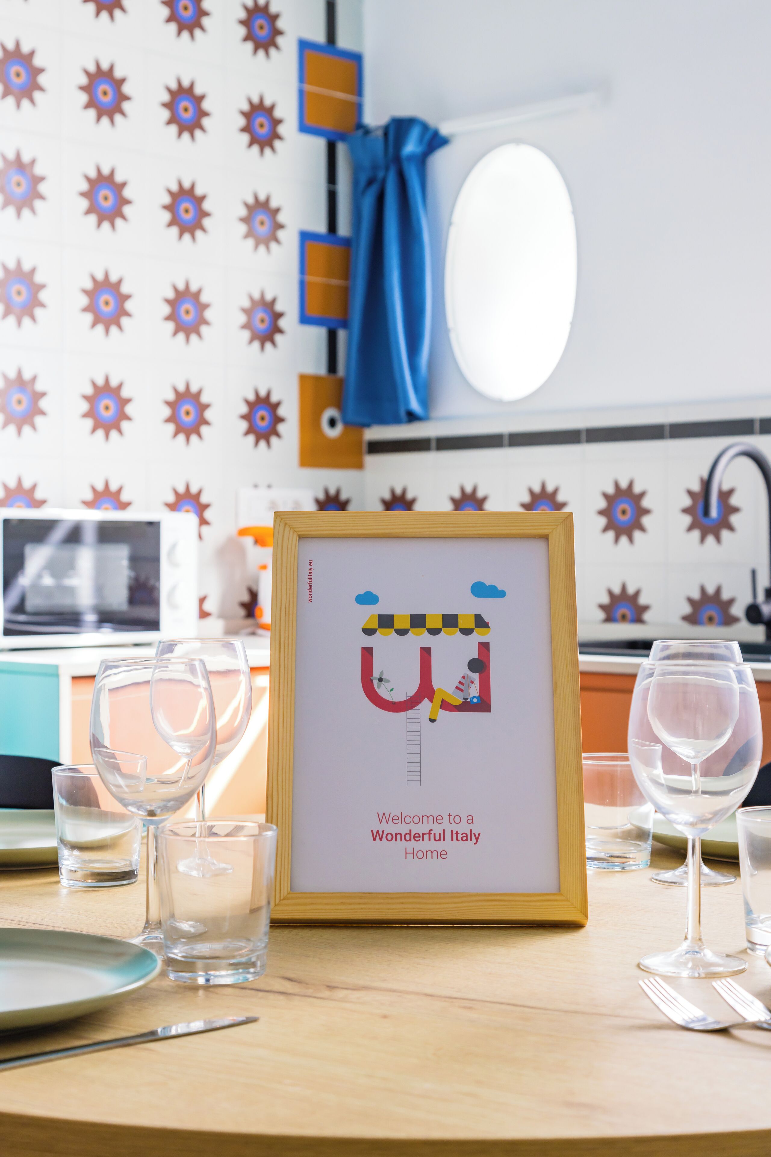 Un cartel de bienvenida en marco con un personaje de caricatura y texto, vasos y cubiertos sobre una mesa, y una zona de cocina con un lavamanos y una toalla azul.