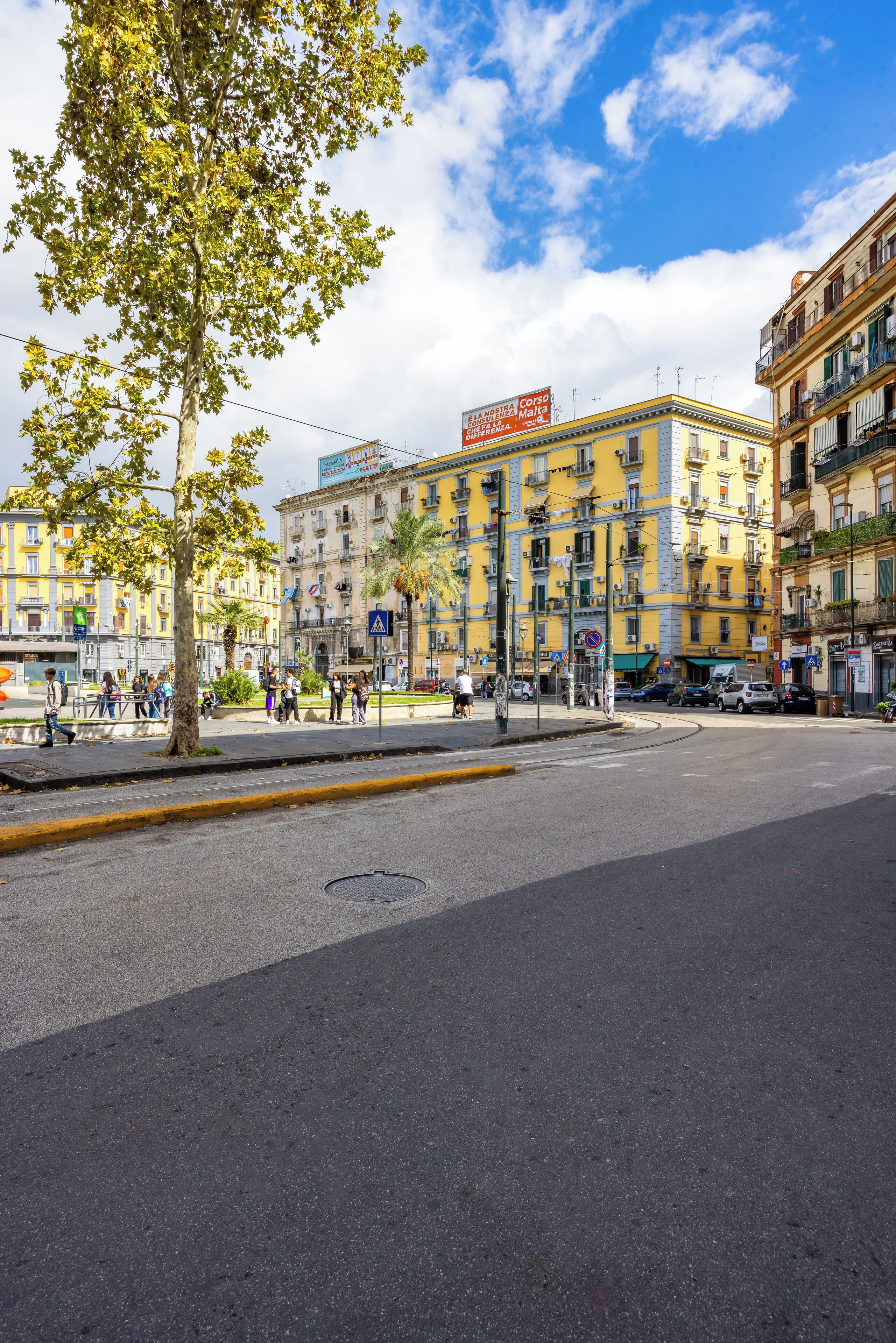 Une scène de rue avec un trottoir bordé d’arbres, des immeubles dotés de balcons et des piétons.